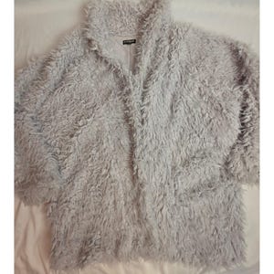 Könnte beinhalten: Eine hellgraue Jacke mit langen Ärmeln und offener Vorderseite aus einem zotteligen, kunstfellartigen Material. Das Kleidungsstück verfügt über einen breiten Kragen und ein glattes, einfarbiges Innenfutter. Ein schwarzes Etikett mit dem weißen Text EXPRESS ist am Hals sichtbar.