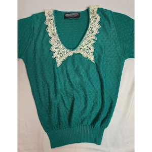 Puede incluir: Una blusa de punto color verde azulado de manga corta con un patrón texturizado de ondas en toda la tela. La prenda tiene un escote en V profundo adornado con encaje color crema y un dobladillo acanalado. Una etiqueta negra dice DAVID BRETT.