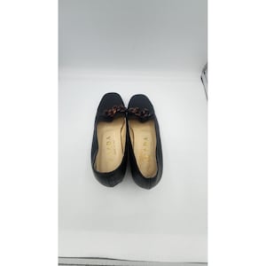Puede incluir: Un par de mocasines de cuero negro con un detalle de cadena con patrón de carey oscuro en el empeine. Las plantillas son de color beige claro y presentan el nombre de la marca Prada en dorado y el texto Made in Italy.