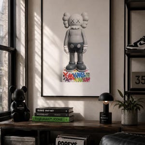 May include: A framed wall art print featuring a gray KAWS character figure standing on a white base covered in colorful graffiti-style text. The figure has white gloved hands, round ears, and X symbols for eyes, centered on a plain white background.