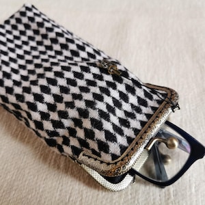 Peut inclure: Un étui à lunettes rectangulaire en tissu à motif losanges noir et blanc. L'étui possède une armature en métal laitonné de style ancien avec un motif gaufré ornemental et une fermeture à fermoir. Une paire de lunettes noires est insérée.