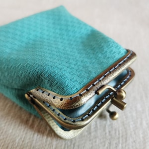 Op de afbeelding: Een kleine tealkleurige portemonnee met een textiel buitenkant voorzien van een ruitpatroon. De portemonnee wordt gesloten door een antiek messingkleurig metalen frame met een knipsluiting en een gedetailleerd bloemmotief in reliëf.