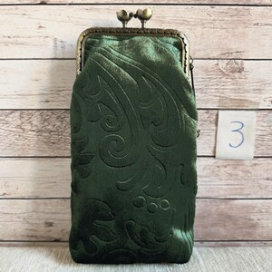 Puede incluir: Un estuche rectangular de terciopelo verde oscuro con un patr&oacute;n floral en relieve y un cierre de marco de metal color bronce con broche de presi&oacute;n en la parte superior. El estuche tiene una superficie suave y texturizada.