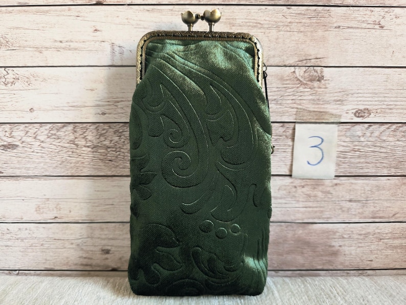 Puede incluir: Un estuche rectangular de terciopelo verde oscuro con un patr&oacute;n floral en relieve y un cierre de marco de metal color bronce con broche de presi&oacute;n en la parte superior. El estuche tiene una superficie suave y texturizada.