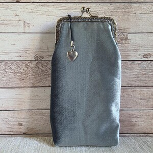 Puede incluir: Un estuche rectangular de terciopelo gris plateado con un cierre met&aacute;lico de boquilla. El estuche presenta un peque&ntilde;o dije plateado en forma de coraz&oacute;n unido al marco mediante un cord&oacute;n negro fino. La tela tiene un brillo suave y uniforme.