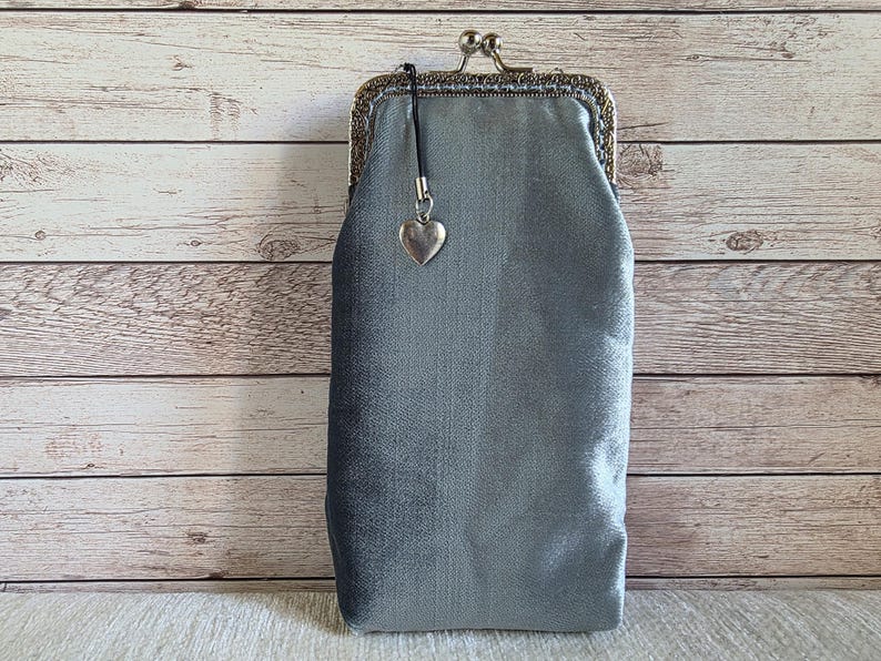 Puede incluir: Un estuche rectangular de terciopelo gris plateado con un cierre met&aacute;lico de boquilla. El estuche presenta un peque&ntilde;o dije plateado en forma de coraz&oacute;n unido al marco mediante un cord&oacute;n negro fino. La tela tiene un brillo suave y uniforme.