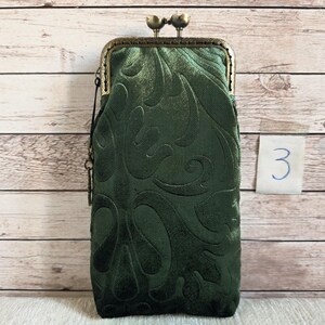 Puede incluir: Un estuche rectangular alto de color verde oscuro con un cierre de broche met&aacute;lico en tono bronce. La tela presenta un patr&oacute;n de remolinos en relieve por toda la superficie. Una peque&ntilde;a etiqueta blanca con el n&uacute;mero 3 escrito a mano est&aacute; en el lateral.