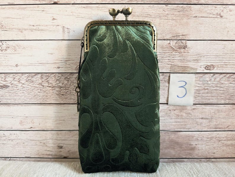 Puede incluir: Un estuche rectangular alto de color verde oscuro con un cierre de broche met&aacute;lico en tono bronce. La tela presenta un patr&oacute;n de remolinos en relieve por toda la superficie. Una peque&ntilde;a etiqueta blanca con el n&uacute;mero 3 escrito a mano est&aacute; en el lateral.