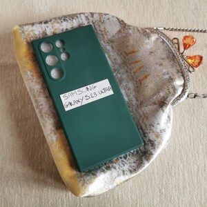 May include: A dark green Samsung Galaxy S23 Ultra phone case with cutouts for the camera lenses, featuring a white rectangular label with the handwritten text SAMSUNG GALAXY S23 ULTRA, resting on a patterned fabric pouch with a silver metal clasp.
