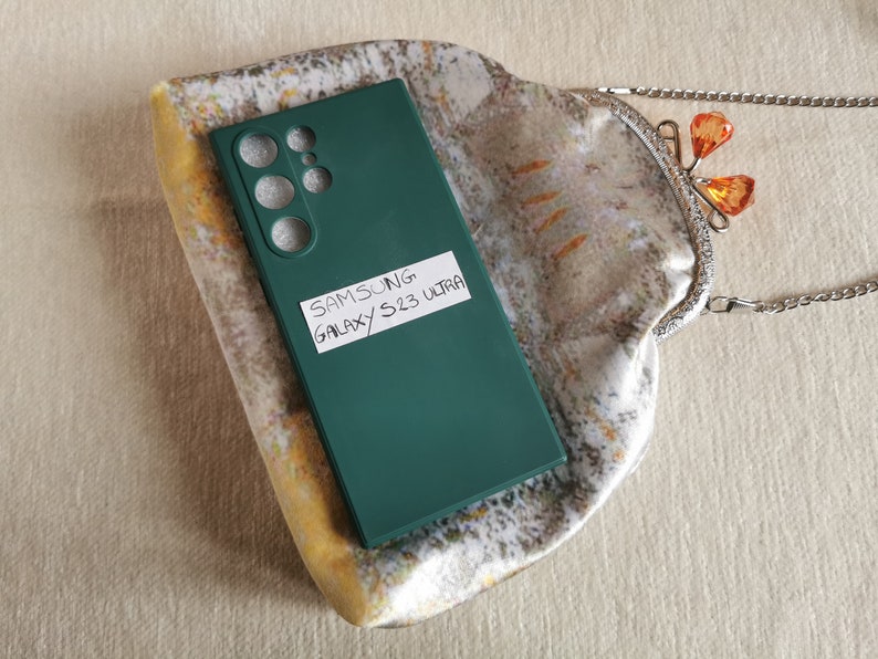 May include: A dark green Samsung Galaxy S23 Ultra phone case with cutouts for the camera lenses, featuring a white rectangular label with the handwritten text SAMSUNG GALAXY S23 ULTRA, resting on a patterned fabric pouch with a silver metal clasp.