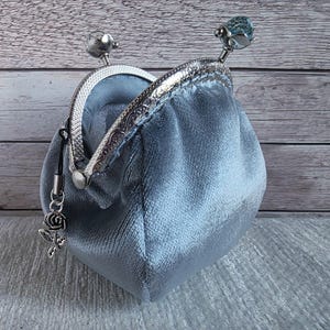 May include: A light blue velvet coin purse with a silver-toned metal kiss-lock frame. The frame features two decorative knobs on top, one of which is a faceted blue bead. A small silver-coloured rose charm hangs from the side of the metal frame.