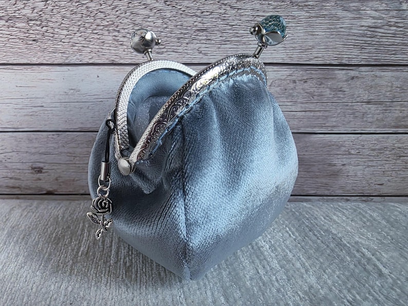 May include: A light blue velvet coin purse with a silver-toned metal kiss-lock frame. The frame features two decorative knobs on top, one of which is a faceted blue bead. A small silver-coloured rose charm hangs from the side of the metal frame.