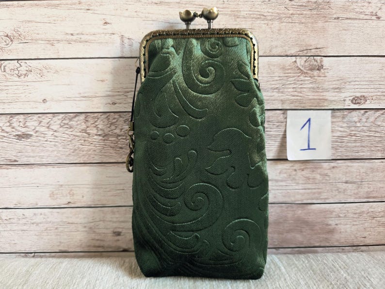 Puede incluir: Un bolso rectangular vertical de tela verde bosque profundo con un patr&oacute;n floral en relieve. El bolso cuenta con un marco de metal color bronce con un cierre de boquilla de dos botones y una peque&ntilde;a anilla lateral para colgar.