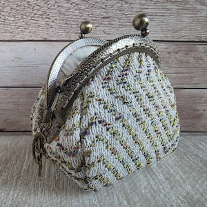 May include: A small coin purse featuring a textured, chevron-patterned fabric in white, yellow, and purple tones. The purse has an antique bronze-coloured metal frame with a kiss-lock closure and a small decorative metal charm attached to the side.