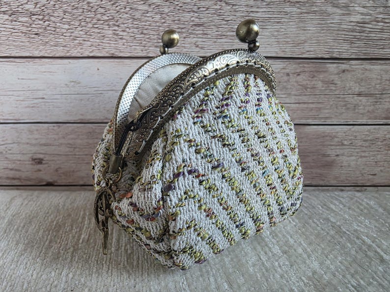 May include: A small coin purse featuring a textured, chevron-patterned fabric in white, yellow, and purple tones. The purse has an antique bronze-coloured metal frame with a kiss-lock closure and a small decorative metal charm attached to the side.