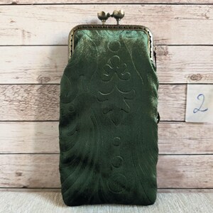 Puede incluir: Un estuche rectangular de terciopelo verde oscuro con un sutil patr&oacute;n floral en relieve y un cierre met&aacute;lico de presi&oacute;n en tono bronce en la parte superior. El estuche presenta una superficie de tela suave y texturizada con un marco r&iacute;gido.