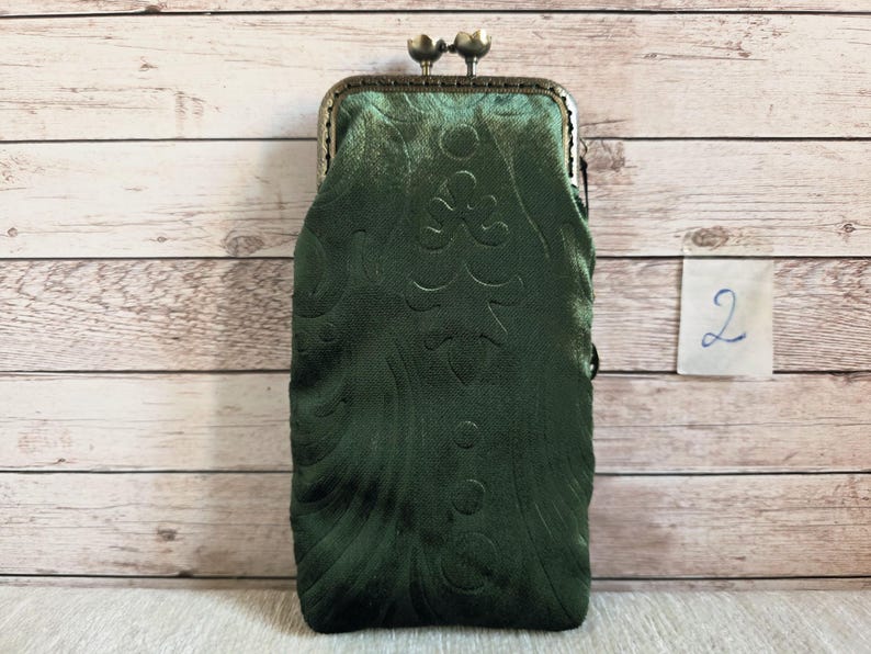 Puede incluir: Un estuche rectangular de terciopelo verde oscuro con un sutil patr&oacute;n floral en relieve y un cierre met&aacute;lico de presi&oacute;n en tono bronce en la parte superior. El estuche presenta una superficie de tela suave y texturizada con un marco r&iacute;gido.