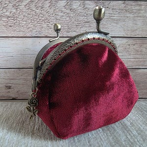 May include: A deep burgundy velvet coin purse featuring an ornate antique bronze metal frame with a kiss-lock closure. The frame is decorated with intricate embossed patterns and includes a small dangling rose charm attached to the side hinge.