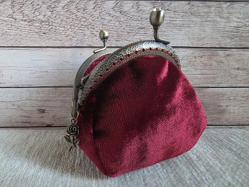 May include: A deep burgundy velvet coin purse featuring an ornate antique bronze metal frame with a kiss-lock closure. The frame is decorated with intricate embossed patterns and includes a small dangling rose charm attached to the side hinge.
