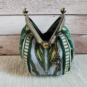 May include: A small coin purse featuring a green and white woven fabric with a repeating pattern. The purse has an antique bronze-coloured metal kiss-lock frame and a small dangling rose-shaped charm attached to the side of the clasp mechanism.