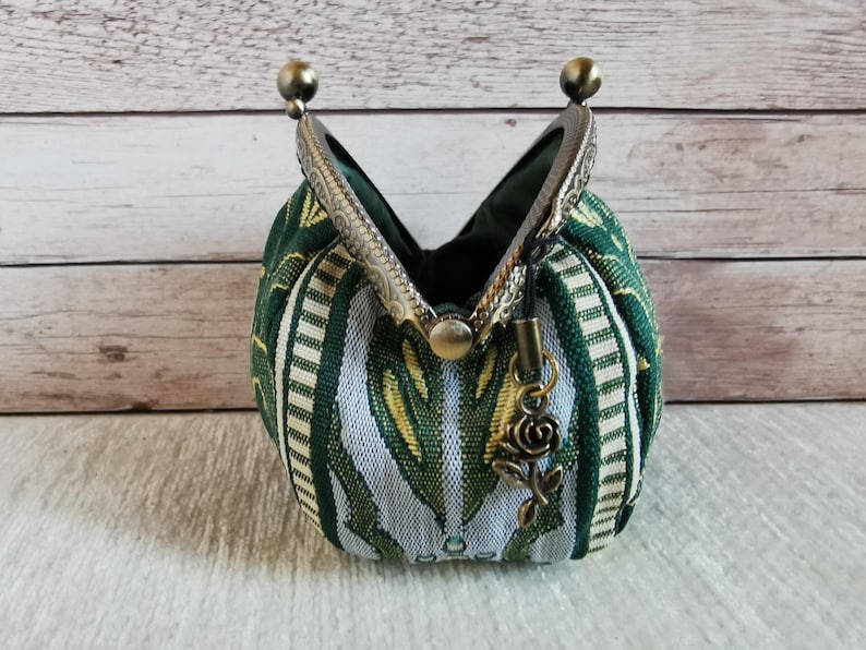 May include: A small coin purse featuring a green and white woven fabric with a repeating pattern. The purse has an antique bronze-coloured metal kiss-lock frame and a small dangling rose-shaped charm attached to the side of the clasp mechanism.