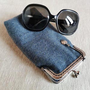 Puede incluir: Un par de gafas de sol negras de gran tama&ntilde;o descansan sobre un estuche rectangular de fieltro azul. El estuche cuenta con un marco de metal plateado con cierre de boquilla y un peque&ntilde;o dije de coraz&oacute;n plateado colgando del cierre.