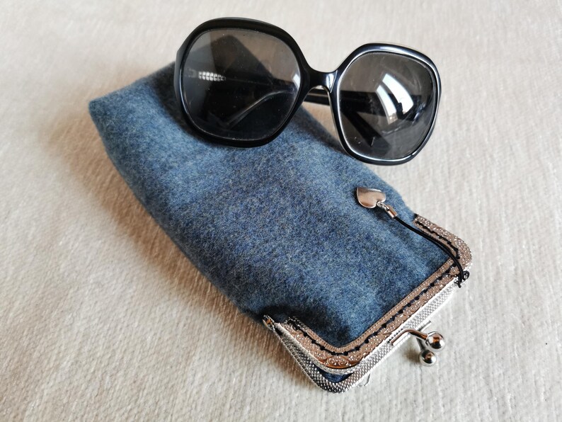 Puede incluir: Un par de gafas de sol negras de gran tama&ntilde;o descansan sobre un estuche rectangular de fieltro azul. El estuche cuenta con un marco de metal plateado con cierre de boquilla y un peque&ntilde;o dije de coraz&oacute;n plateado colgando del cierre.