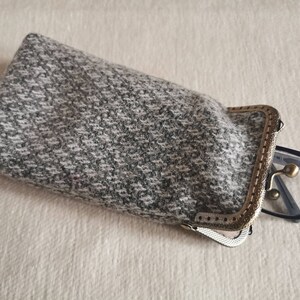 Puede incluir: Un estuche rectangular para gafas hecho de tela de tweed moteada en gris y blanco. El estuche cuenta con un marco de metal en tono lat&oacute;n antiguo con cierre de boquilla. Unas gafas de montura negra asoman parcialmente por la abertura.