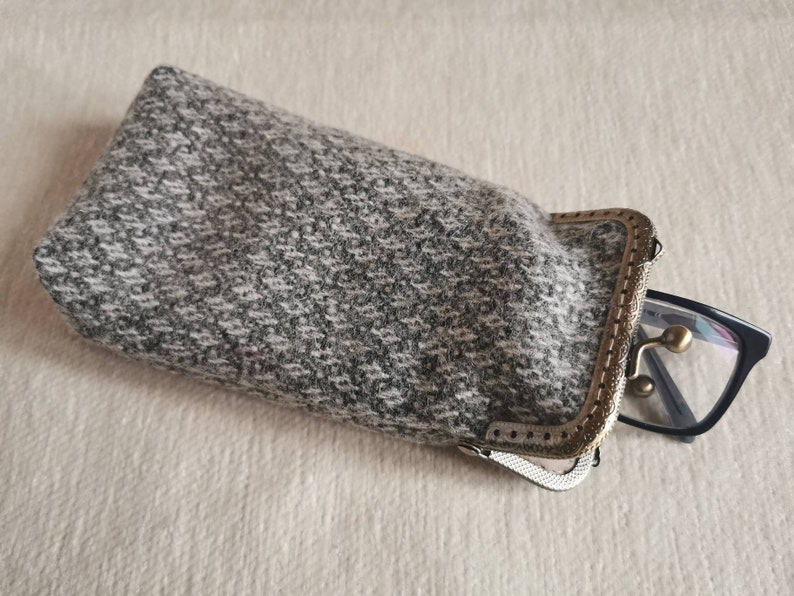 Puede incluir: Un estuche rectangular para gafas hecho de tela de tweed moteada en gris y blanco. El estuche cuenta con un marco de metal en tono lat&oacute;n antiguo con cierre de boquilla. Unas gafas de montura negra asoman parcialmente por la abertura.