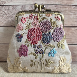 May include: A small coin purse with a cream-coloured fabric base featuring an intricate floral embroidery design in shades of red, purple, pink, and blue. The purse has a metallic bronze-toned kiss-lock clasp closure and decorative gold-toned lace trim.
