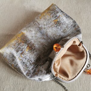 May include: A clutch bag featuring a textured grey and white fabric with gold accents. The bag has a silver-toned metal frame with a kiss-lock closure topped with two orange faceted beads. It includes a silver chain strap and a beige interior lining.
