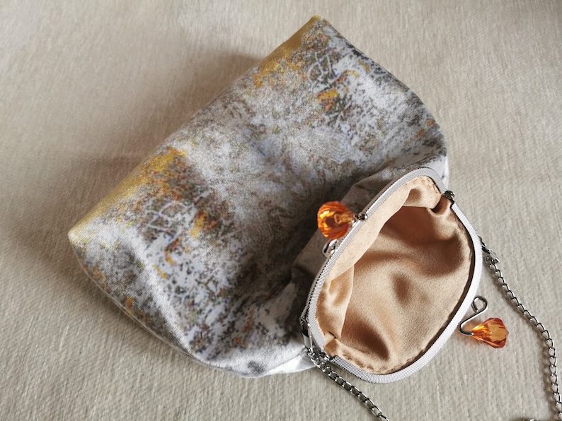 May include: A clutch bag featuring a textured grey and white fabric with gold accents. The bag has a silver-toned metal frame with a kiss-lock closure topped with two orange faceted beads. It includes a silver chain strap and a beige interior lining.