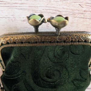 Puede incluir: Un monedero de terciopelo verde oscuro con un patr&oacute;n de remolinos en relieve. El monedero tiene un marco de metal color bronce con detalles de filigrana y un cierre de boquilla rematado con dos cuentas verde p&aacute;lido en forma de flor.