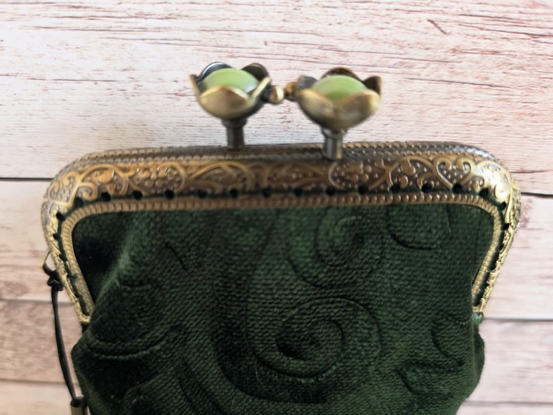 Puede incluir: Un monedero de terciopelo verde oscuro con un patr&oacute;n de remolinos en relieve. El monedero tiene un marco de metal color bronce con detalles de filigrana y un cierre de boquilla rematado con dos cuentas verde p&aacute;lido en forma de flor.