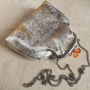 May include: A silver and gold metallic fabric clutch bag with a kiss-lock frame closure. The bag features a detachable silver-tone metal chain strap and two faceted orange crystal charms attached to the clasp mechanism of the frame.