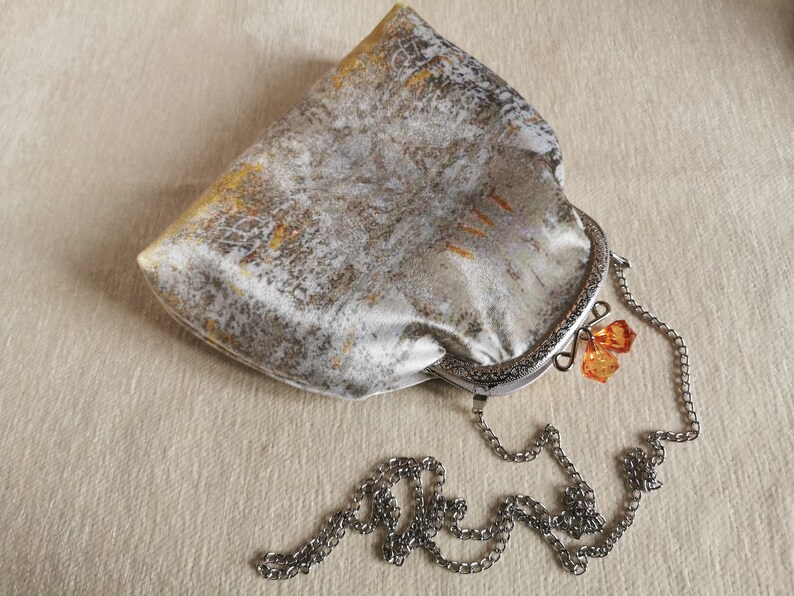 May include: A silver and gold metallic fabric clutch bag with a kiss-lock frame closure. The bag features a detachable silver-tone metal chain strap and two faceted orange crystal charms attached to the clasp mechanism of the frame.