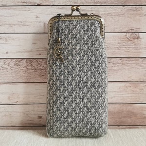 Puede incluir: Un estuche rectangular de tela de tweed gris y blanca con un patr&oacute;n de diamantes texturizado. Cuenta con un cierre de boquilla met&aacute;lico en tono bronce y un peque&ntilde;o dije colgante con forma de media luna con una figura sentada.