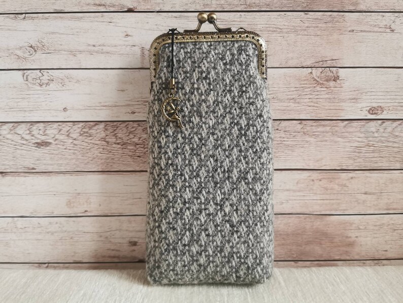 Puede incluir: Un estuche rectangular de tela de tweed gris y blanca con un patr&oacute;n de diamantes texturizado. Cuenta con un cierre de boquilla met&aacute;lico en tono bronce y un peque&ntilde;o dije colgante con forma de media luna con una figura sentada.