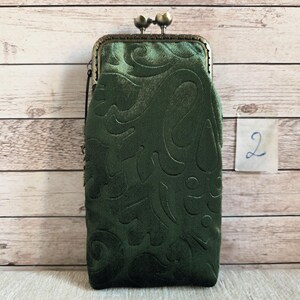 Puede incluir: Un bolso rectangular vertical de terciopelo verde oscuro con un patr&oacute;n floral en relieve. El bolso cuenta con un cierre de marco met&aacute;lico de color bronce con dos pomos esf&eacute;ricos en la parte superior y una etiqueta con el n&uacute;mero 2.
