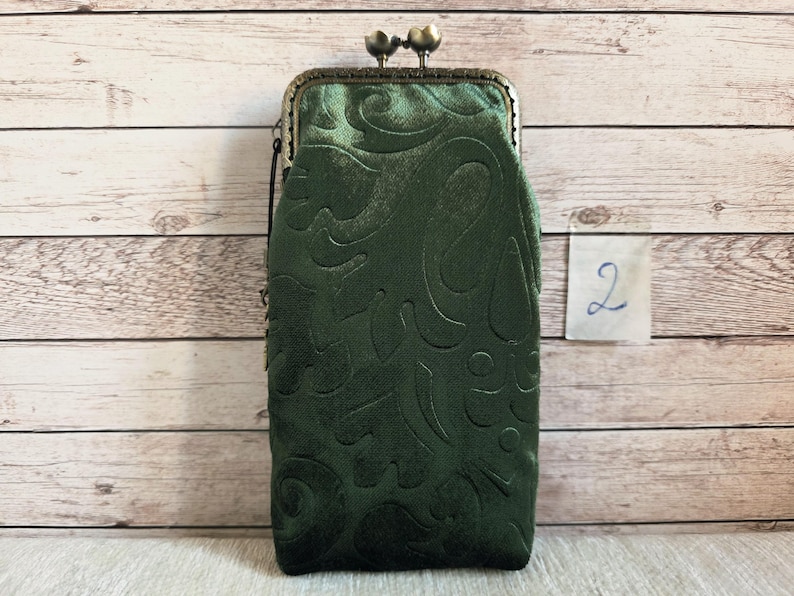 Puede incluir: Un bolso rectangular vertical de terciopelo verde oscuro con un patr&oacute;n floral en relieve. El bolso cuenta con un cierre de marco met&aacute;lico de color bronce con dos pomos esf&eacute;ricos en la parte superior y una etiqueta con el n&uacute;mero 2.