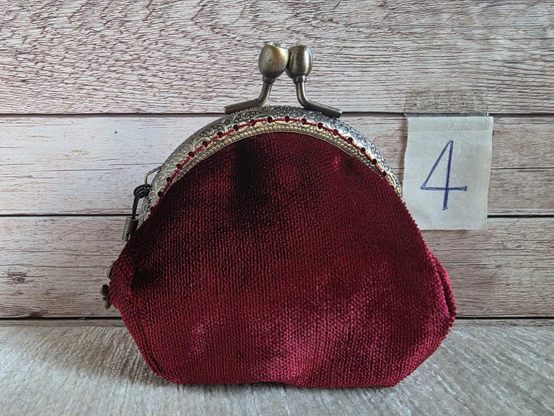 May include: A deep red velvet coin purse with a textured, antique bronze-toned metal frame and a dual-ball clasp closure. A small white paper tag with the handwritten number 4 is attached to the side of the frame, featuring a decorative edge.
