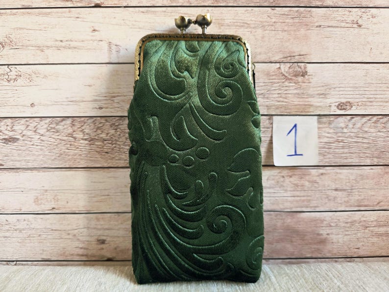 Puede incluir: Un estuche rectangular de color verde oscuro con un patr&oacute;n floral en relieve. El estuche tiene un marco met&aacute;lico de tono bronce con un cierre de doble bola en la parte superior. Se observa una peque&ntilde;a etiqueta cuadrada blanca con el n&uacute;mero 1.