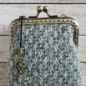 Puede incluir: Un monedero de tweed gris y blanco con un patr&oacute;n de diamantes texturizado, un marco de metal color bronce con cierre de boquilla y detalles de filigrana, y un peque&ntilde;o dije de bronce de una hada sentada en una luna creciente.