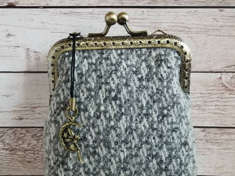 Puede incluir: Un monedero de tweed gris y blanco con un patr&oacute;n de diamantes texturizado, un marco de metal color bronce con cierre de boquilla y detalles de filigrana, y un peque&ntilde;o dije de bronce de una hada sentada en una luna creciente.