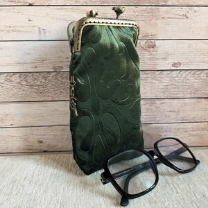 Puede incluir: Un estuche para gafas de terciopelo verde oscuro con un patr&oacute;n en relieve, que presenta un cierre de boquilla de metal color bronce y un peque&ntilde;o dije colgante en el lateral. Unas gafas de montura negra descansan frente al estuche vertical.