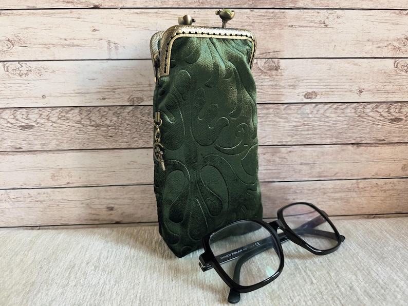 Puede incluir: Un estuche para gafas de terciopelo verde oscuro con un patr&oacute;n en relieve, que presenta un cierre de boquilla de metal color bronce y un peque&ntilde;o dije colgante en el lateral. Unas gafas de montura negra descansan frente al estuche vertical.