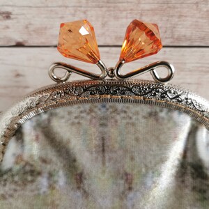 May include: A coin purse featuring a silver-toned metal frame with an intricate embossed pattern and two faceted orange crystal-like beads as a clasp. The body of the purse is made of a soft fabric with a muted, mottled pattern in shades of green and grey.