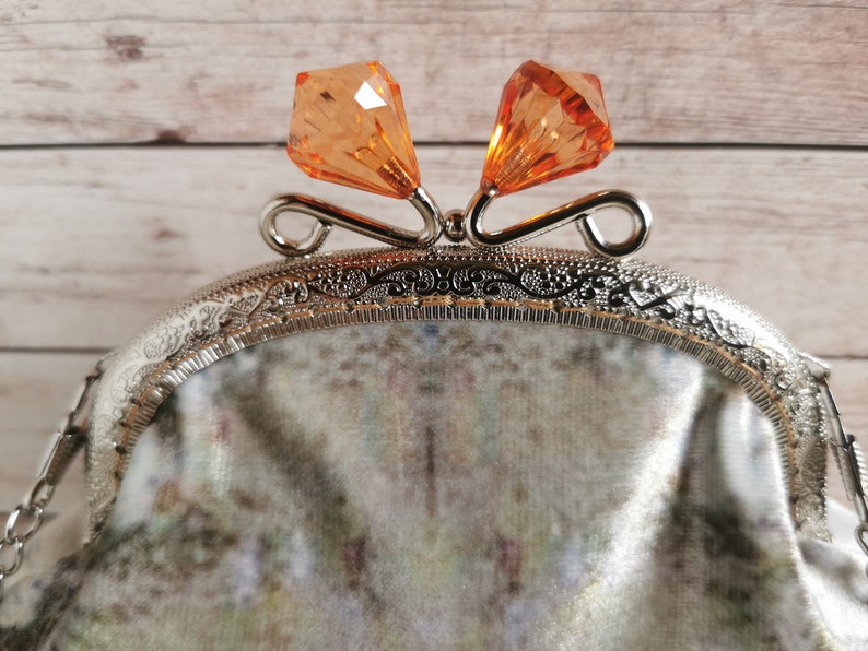 May include: A coin purse featuring a silver-toned metal frame with an intricate embossed pattern and two faceted orange crystal-like beads as a clasp. The body of the purse is made of a soft fabric with a muted, mottled pattern in shades of green and grey.