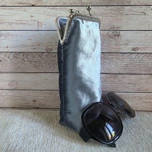 Puede incluir: Un estuche vertical de terciopelo gris plateado con un cierre met&aacute;lico de boquilla. El estuche est&aacute; colocado en posici&oacute;n vertical junto a unas gafas de sol con montura negra y cristales oscuros. El terciopelo tiene un brillo suave.
