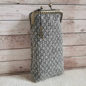 Puede incluir: Un estuche rectangular hecho de tela de tweed gris y blanca con un patr&oacute;n de diamantes texturizado. El estuche cuenta con un cierre de marco de metal color lat&oacute;n antiguo y un peque&ntilde;o dije colgante con forma de luna creciente y estrella.